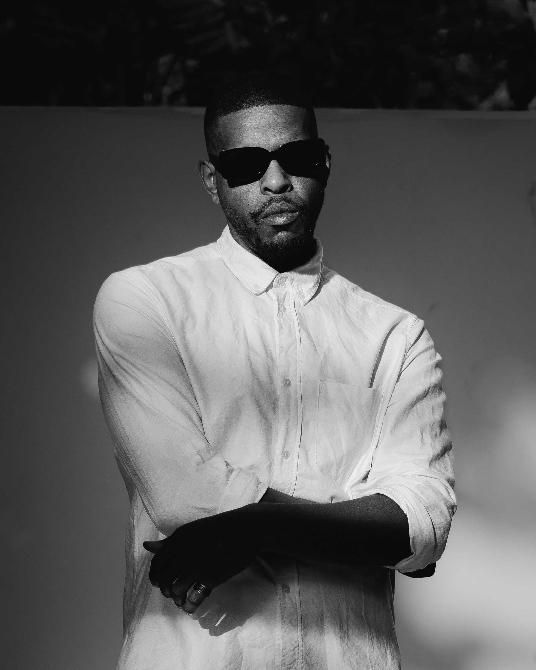 Black and white portrait of Scotty Grand in a white shirt and dark sunglasses, arms folded, against a sunlit wall